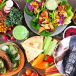 Uma variedade diversificada de itens de almoço saudáveis, incluindo uma tigela de Buda, wraps de alface, sanduíches, salada, homus, vegetais e um smoothie verde em um fundo de madeira.