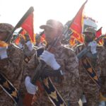 Militares do IRGC marchando em desfile, segurando rifles e bandeiras vermelhas.