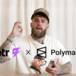 A Betr de Jake Paul faz parceria com a Polymarket para lançar mercados de previsão dentro do aplicativo. Jake Paul está sentado em uma mesa segurando um telefone enquanto os logotipos Betr e Polymarket aparecem na tela, anunciando uma parceria entre o aplicativo de apostas e a plataforma de mercado de previsão.