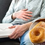 Homem de jeans e moletom cinza esfregando a barriga enquanto segura uma caixa de comida branca com um garfo de plástico no colo.