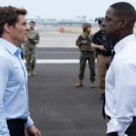 Two men stand opposite each other in shirts on an airport runway