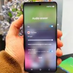 Um smartphone Galaxy S26 cercado por emojis raivosos e símbolos de fogo, sobrepostos com um carimbo vermelho 'CALM DOWN'