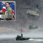 Um helicóptero militar carregando um grande contêiner sobrevoa um rio onde soldados em um barco com bandeira americana observam.