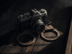 Escoteiro de modelos infantis de Michigan acusado de apostar milhões em caso de fraude Escoteiro de modelos infantis de Michigan acusado de apostar milhões em casos de fraude. Uma câmera de filme vintage apoiada em uma superfície de madeira, posicionada entre um par de algemas de metal sob uma iluminação fraca e dramática.