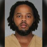 Foto de um homem com cabelos escuros e barba, vestindo um macacão laranja.