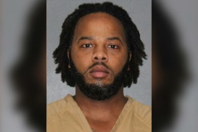 Foto de um homem com cabelos escuros e barba, vestindo um macacão laranja.