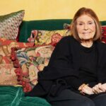 Gloria Steinem sentada em um sofá verde, rodeada de almofadas decorativas, com uma grande gravura emoldurada de uma criança lendo ao lado de um elefante ao fundo.