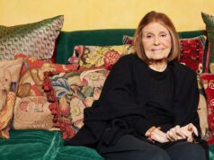 Gloria Steinem leiloando pela primeira vez itens pessoais que colecionou durante sua carreira Gloria Steinem sentada em um sofá verde, rodeada de almofadas decorativas, com uma grande gravura emoldurada de uma criança lendo ao lado de um elefante ao fundo.
