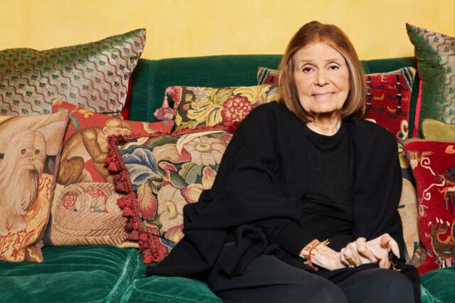 Gloria Steinem sentada em um sofá verde, rodeada de almofadas decorativas, com uma grande gravura emoldurada de uma criança lendo ao lado de um elefante ao fundo.