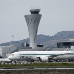 Um jato de passageiros ZipAir taxiando com uma torre de controle de tráfego aéreo e colinas ao fundo.