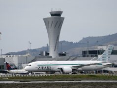 Grandes atrasos esperados no aeroporto de São Francisco sob os novos limites da FAA Um jato de passageiros ZipAir taxiando com uma torre de controle de tráfego aéreo e colinas ao fundo.