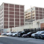 Exterior do Centro de Detenção Metropolitano no Brooklyn, um prédio de vários andares com painéis de tijolos e estrutura de concreto, visto de uma rua com carros estacionados.