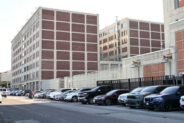 Exterior do Centro de Detenção Metropolitano no Brooklyn, um prédio de vários andares com painéis de tijolos e estrutura de concreto, visto de uma rua com carros estacionados.