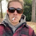 Um homem de flanela fumando um cigarro lá fora.