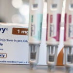 Dr. Sakthi Karunanithi em um estúdio da BBC com um fundo de árvores e casas contra um céu azul. Dr. Sakthi tem cabelo branco penteado para trás e uma barba bem cuidada e está vestindo um paletó cinza, gravata floral verde e camisa listrada