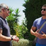 Um close de um homem de óculos escuros e terno, lá fora, ao sol.