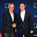 O comissário da MLB, Rob Manfred, e o presidente da CFTC, Mike Selig, apertam as mãos no evento de anúncio de parceria da Liga Principal de Beisebol com logotipos da MLB como pano de fundo