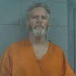 Mugshot de Richard Chesher, um homem com barba e cabelos grisalhos, vestindo um macacão laranja.