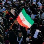 Multidão de mulheres em hijabs e xadores, com uma bandeira iraniana no centro, num comício.