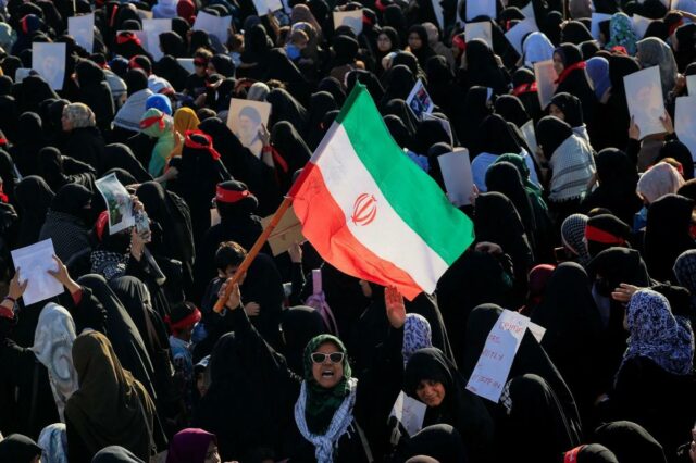 Multidão de mulheres em hijabs e xadores, com uma bandeira iraniana no centro, num comício.