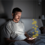 Homem deitado na cama à noite olhando para um smartphone brilhante enquanto ícones estilizados de moedas de ouro flutuam para cima na tela, sugerindo jogos de azar online ou atividades de apostas enquanto outra pessoa dorme ao lado dele em um quarto mal iluminado. O boom das apostas digitais remodela os hábitos de jogo americanos, levantando preocupações sobre danos financeiros