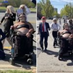 Um manifestante em uma cadeira de rodas, usando uma proteção facial e uma blusa preta, segura uma coleira presa a um homem de chapéu e terno vermelho, semelhante a Donald Trump, em uma rua com outros manifestantes e um carro da polícia.