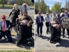 O protesto ‘No Kings’ em Atlanta se torna estranho quando esquerdistas mascarados e usuários de cadeiras de rodas caminham como Trump na coleira Um manifestante em uma cadeira de rodas, usando uma proteção facial e uma blusa preta, segura uma coleira presa a um homem de chapéu e terno vermelho, semelhante a Donald Trump, em uma rua com outros manifestantes e um carro da polícia.