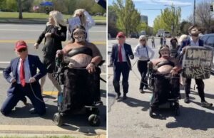 O protesto ‘No Kings’ em Atlanta se torna estranho quando esquerdistas mascarados e usuários de cadeiras de rodas caminham como Trump na coleira Um manifestante em uma cadeira de rodas, usando uma proteção facial e uma blusa preta, segura uma coleira presa a um homem de chapéu e terno vermelho, semelhante a Donald Trump, em uma rua com outros manifestantes e um carro da polícia.