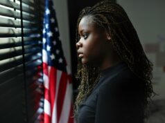 Os jovens americanos são infelizes – apesar dos jovens mais felizes do resto do mundo Retrato de uma jovem adolescente estressada olhando pela janela com uma bandeira americana ao fundo.