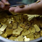 Uma mão coloca uma pequena barra de ouro em um recipiente cheio de várias barras e peças de ouro.