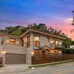 Uma casa de pedra com telhado de terracota, varandas e uma longa cerca de metal estampada na frente, com uma encosta de árvores verdes e arbustos atrás dela.
