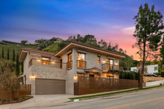Uma casa de pedra com telhado de terracota, varandas e uma longa cerca de metal estampada na frente, com uma encosta de árvores verdes e arbustos atrás dela.