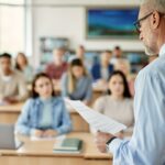 Um professor de cabelos grisalhos e óculos lendo um jornal para uma sala de aula de alunos.