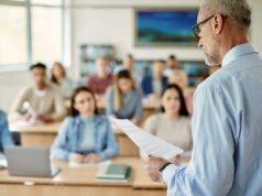 Professor revela o sinal revelador que os alunos estão usando IA em suas tarefas Um professor de cabelos grisalhos e óculos lendo um jornal para uma sala de aula de alunos.