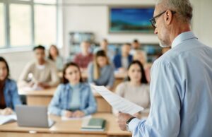 Professor revela o sinal revelador que os alunos estão usando IA em suas tarefas Um professor de cabelos grisalhos e óculos lendo um jornal para uma sala de aula de alunos.