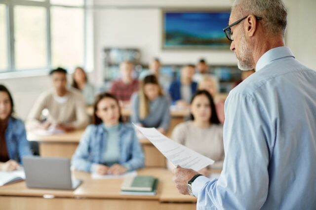 Um professor de cabelos grisalhos e óculos lendo um jornal para uma sala de aula de alunos.