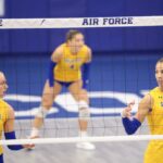 Três jogadores de vôlei com uniformes amarelos e azuis estão em uma quadra atrás de uma rede.