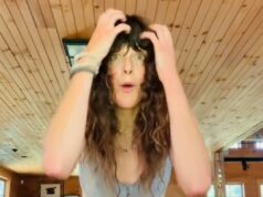 Sacuda isso! Terapia somática, uma maneira maluca e selvagem de vencer o estresse Uma mulher com cabelos castanhos cacheados demonstra tremores somáticos em uma sala com painéis de madeira.