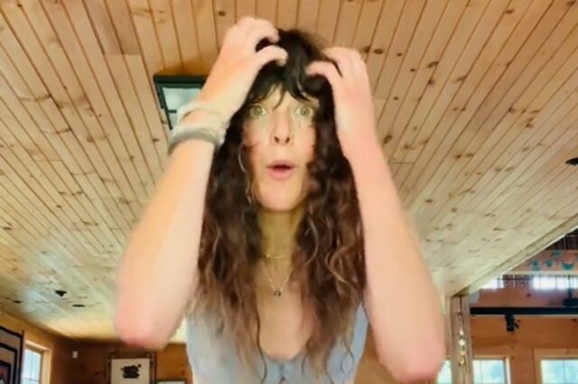 Uma mulher com cabelos castanhos cacheados demonstra tremores somáticos em uma sala com painéis de madeira.