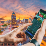 Nova Assembleia de Wisconsin avança plano de apostas esportivas móveis em todo o estado controlado por tribos. Close de uma pessoa segurando um smartphone exibindo um aplicativo de apostas esportivas ao vivo, com o horizonte de uma cidade ao pôr do sol ao fundo, com prédios altos e um rio atravessando o centro da cidade.