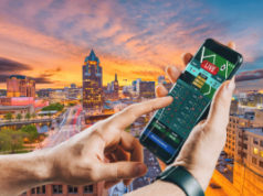 Senado de Wisconsin avança estrutura tribal para expansão de apostas esportivas móveis Nova Assembleia de Wisconsin avança plano de apostas esportivas móveis em todo o estado controlado por tribos. Close de uma pessoa segurando um smartphone exibindo um aplicativo de apostas esportivas ao vivo, com o horizonte de uma cidade ao pôr do sol ao fundo, com prédios altos e um rio atravessando o centro da cidade.