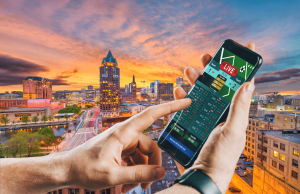 Senado de Wisconsin avança estrutura tribal para expansão de apostas esportivas móveis Nova Assembleia de Wisconsin avança plano de apostas esportivas móveis em todo o estado controlado por tribos. Close de uma pessoa segurando um smartphone exibindo um aplicativo de apostas esportivas ao vivo, com o horizonte de uma cidade ao pôr do sol ao fundo, com prédios altos e um rio atravessando o centro da cidade.