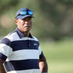 Tiger Woods em uma camisa pólo listrada de azul e branco e boné azul com óculos escuros.