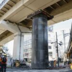 Pessoas com capacetes e coletes se reúnem em torno de um grande cano que emergiu do solo em um canteiro de obras de esgoto sob um viaduto em Osaka, no Japão.
