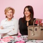 Uma jovem de jeans e camisa azul clara está sentada em um banquinho, segurando aberto um jornal do New York Post com a manchete