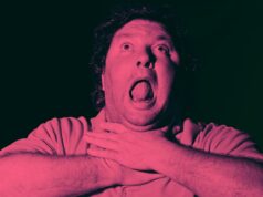 Você vai engasgar ao saber quantos empregos em tempo integral um data center de US$ 136 milhões realmente criará Um homem de cabelos cacheados é mostrado com a boca bem aberta e os olhos voltados para cima, segurando a garganta com as duas mãos, como se estivesse sufocando ou lutando para respirar. A imagem tem uma forte tonalidade vermelho-rosada e um fundo escuro.