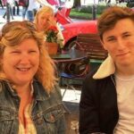 Um jovem dormindo em sua cama, abraçando seu cachorro. O homem veste uma camisa de futebol amarela e está com o braço esquerdo sobre o cachorro. O cachorro tem pelo preto.