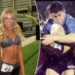 Uma líder de torcida com longos cabelos loiros e vestindo uma blusa preta brilhante e shorts com o número 45 sorri para a câmera em um campo de futebol.