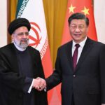 O presidente iraniano, Ebrahim Raisi, e o presidente chinês, Xi Jinping, apertam as mãos diante de suas bandeiras nacionais.