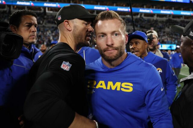 Dois homens, um com capuz azul e outro com camiseta escura, sentam-se em uma mesa com microfones em frente a um fundo azul com 